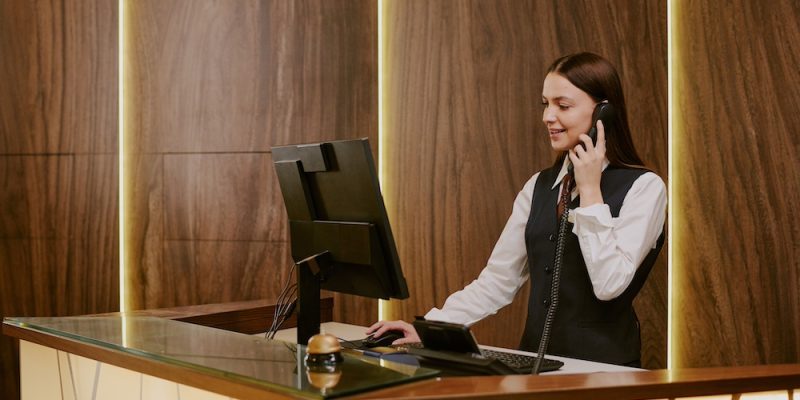 Hotel Receptionist Answering Call Wide shot of young adult woman looking at computers screen and answering phone call while working as receptionist at hotel